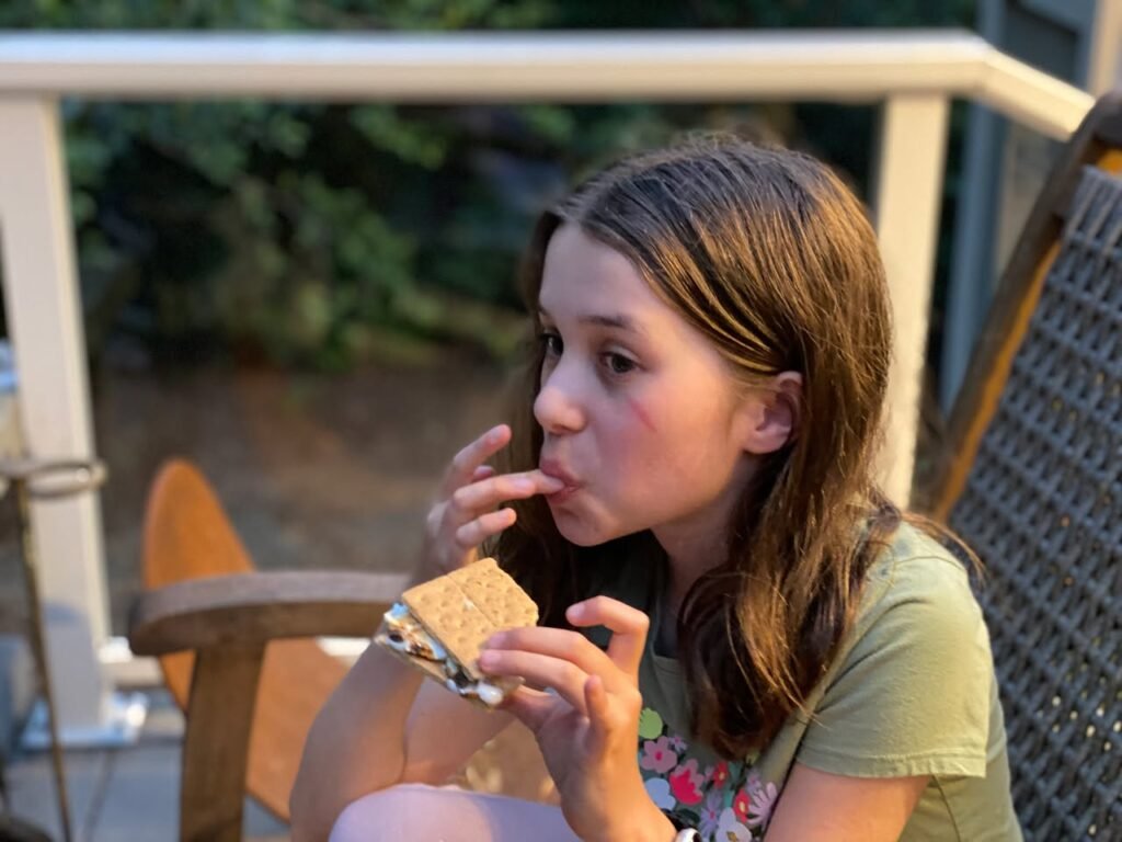 A person seated on a back deck holding a sandwich on a paper napkin. A close-up view of hair against a blurred outdoor backdrop.