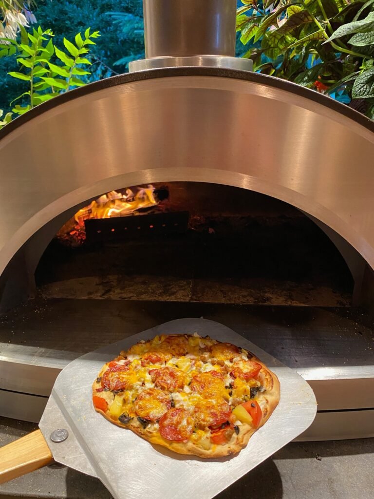 A freshly cooked pizza topped with melted cheese and vegetables is being removed from a stainless steel wood-fired outdoor oven. Lush green foliage surrounds the cooking area.