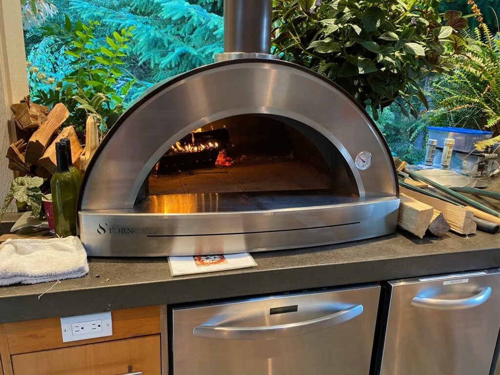 A stainless steel wood-fired pizza oven on a patio counter surrounded by greenery and firewood.