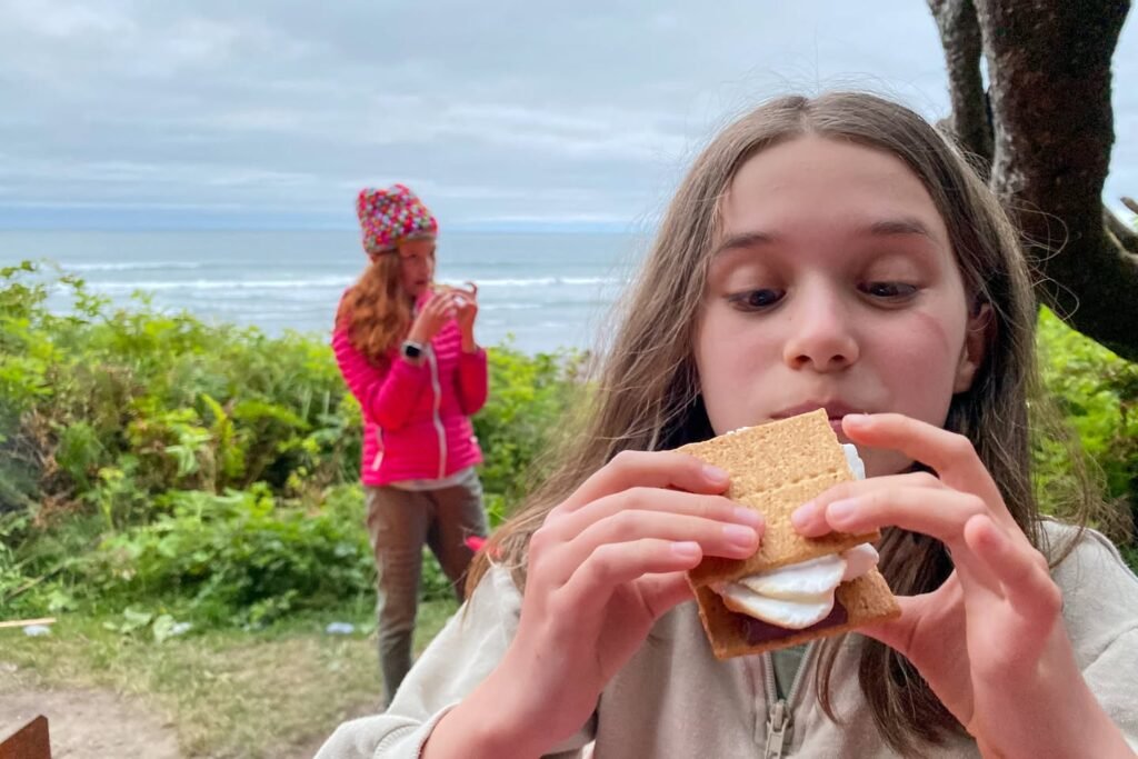 A close-up of someone eating a s'more with an ocean and greenery in the background, while another person stands in the distance.