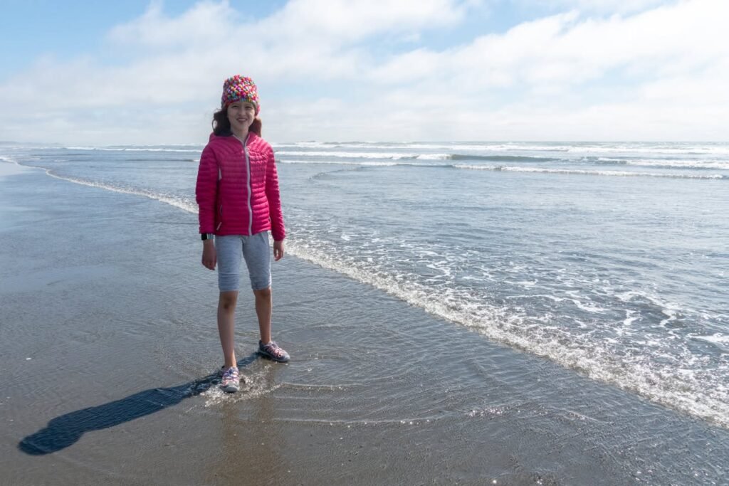 A person in a red jacket stands on a sandy beach with gentle waves lapping at their feet. The wide shot features a vast, calm ocean under a blue sky.