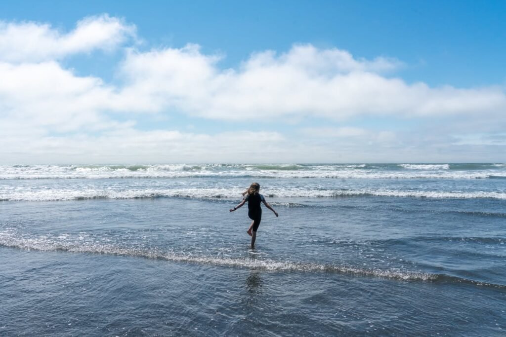 A child runs happily through shallow waves on a sunny beach, with a bright blue sky and clouds overhead.
