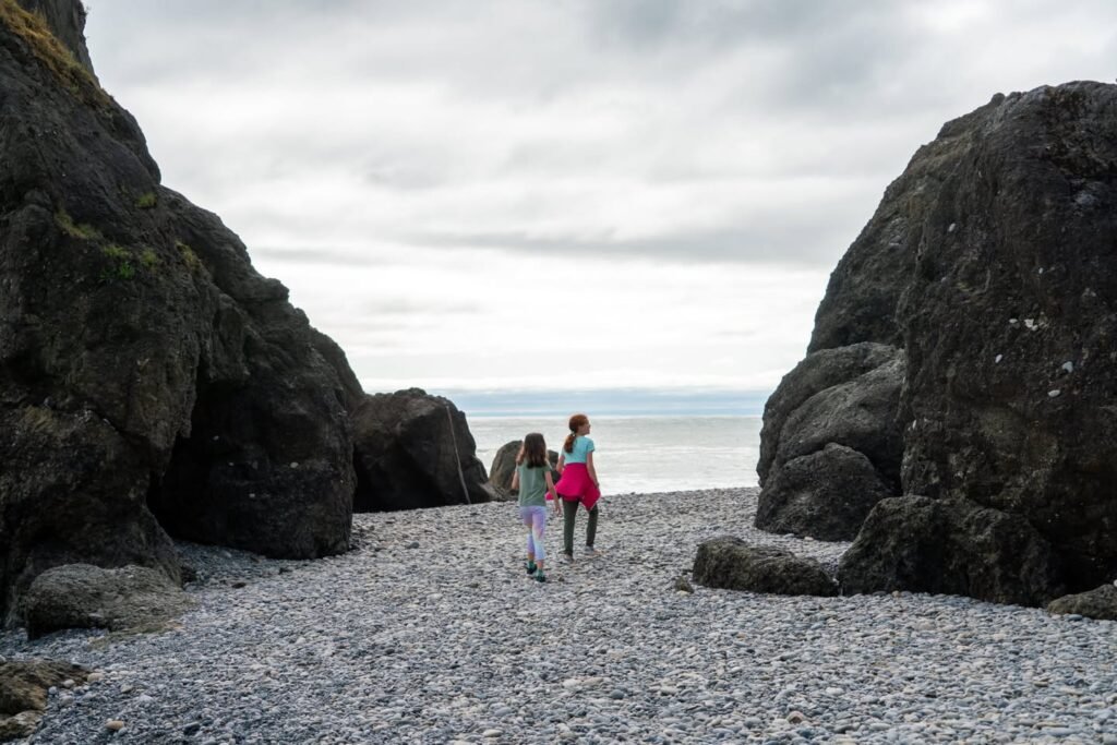 Two figures walk along a rocky beach path between massive sea stacks towards the horizon.