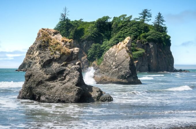 A foggy view of a Pacific Northwest rocky island surrounded by turquoise waves. Giant rock formations are partially visible in the mist.