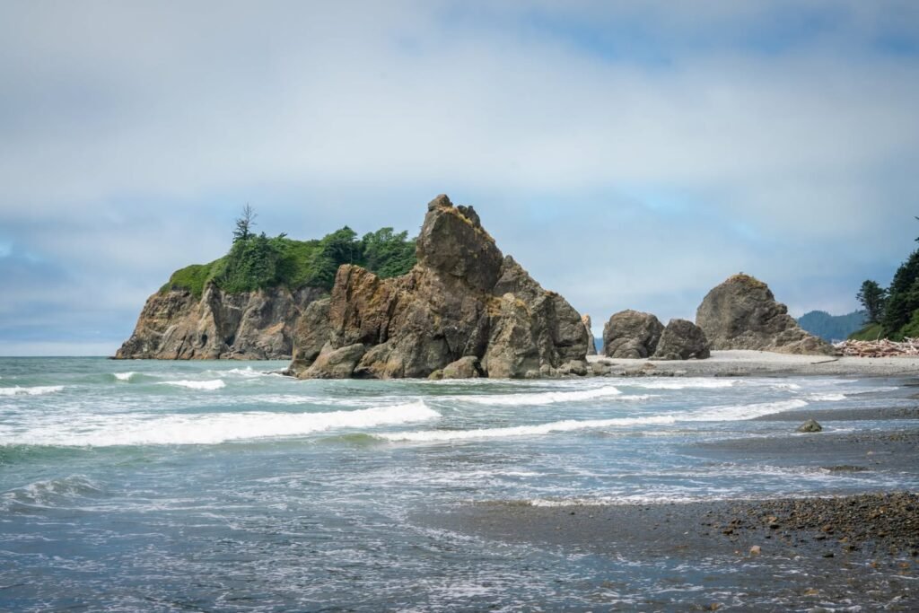 Rocky coastal outcrops rise from the ocean beneath a partly cloudy sky. Gentle waves wash against the shore near rugged cliffs.