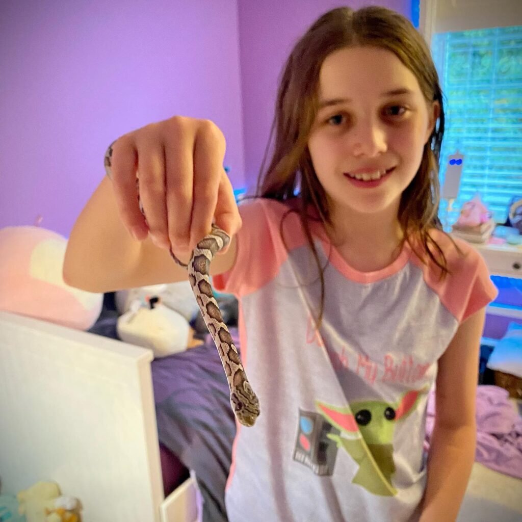 A smiling young person holds a patterned snake in a bedroom with pink wallpaper.