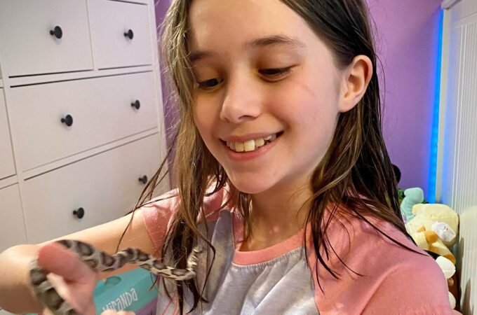 A person smiles happily while holding a small, patterned snake. The scene takes place in a bedroom with purple walls.