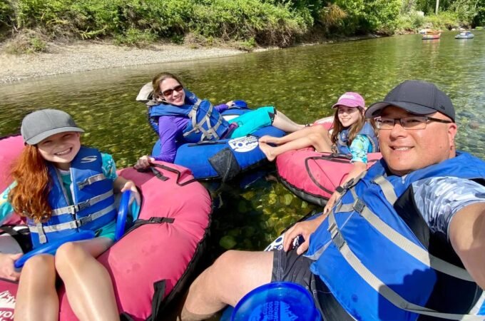 A family enjoying a relaxing river floating trip in colorful inflatable tubes, surrounded by lush greenery. A self-portrait captures the group in safe life jackets on calm waters.