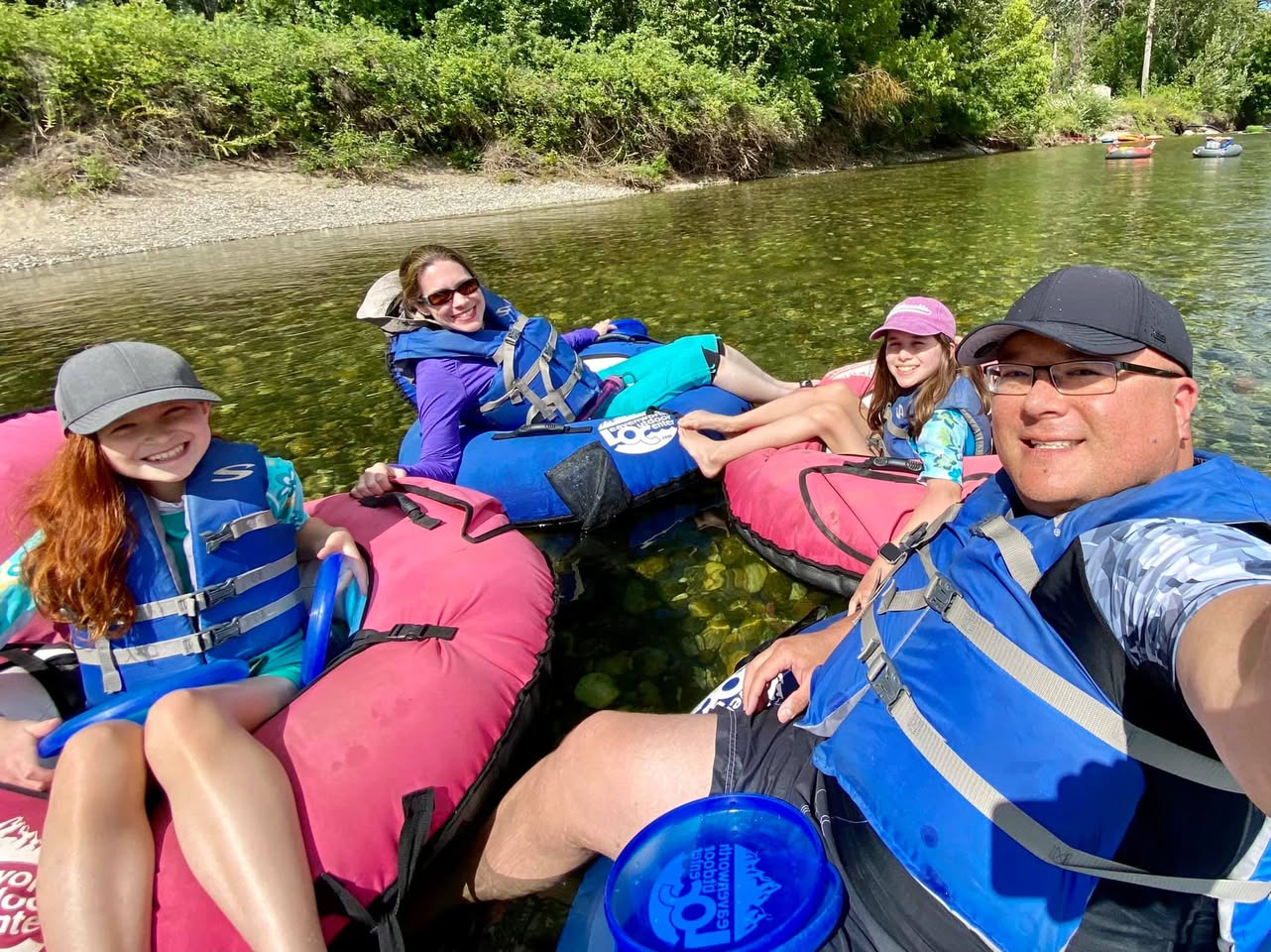 A family enjoying a relaxing river floating trip in colorful inflatable tubes, surrounded by lush greenery. A self-portrait captures the group in safe life jackets on calm waters.