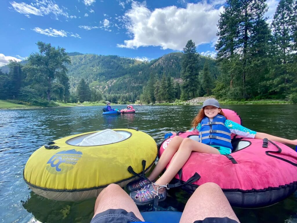 A person relaxes on a pink inflatable tube on a calm river surrounded by lush green mountains and forest. Other tubes and kayakers are visible in the background under a bright blue sky.