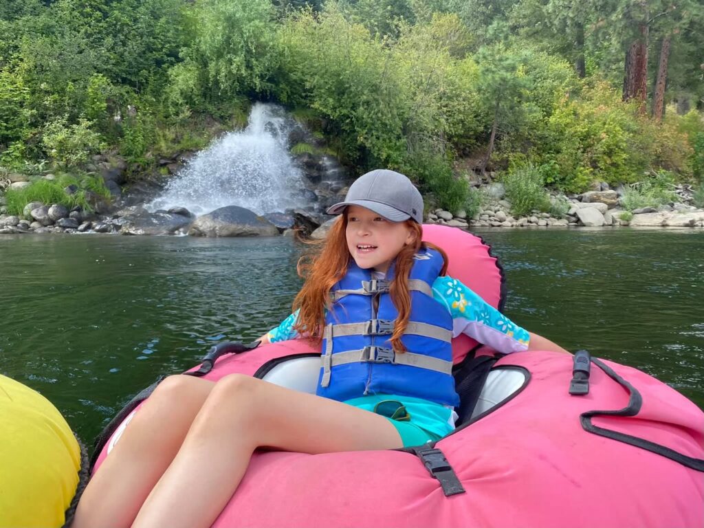 A child floats on a pink inflatable raft, wearing a life vest and cap, with lush greenery and a waterfall in the background.