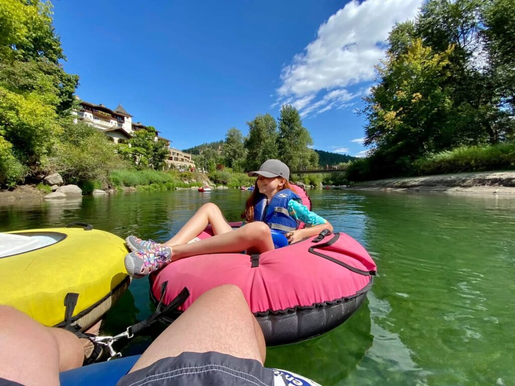 People relax on inflatable tubes on a calm, green river surrounded by lush vegetation under a bright blue sky.