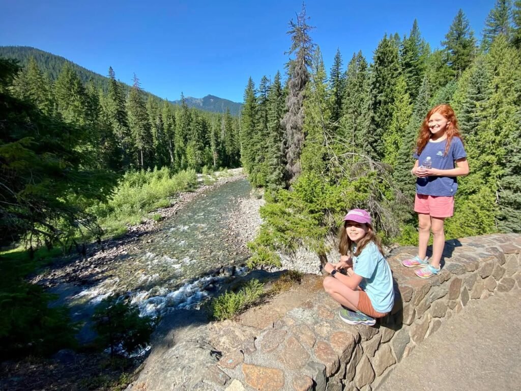 Two individuals enjoy a scenic overlook with a river and evergreen forest in the background. A close-up of the lush mountain landscape highlights the dense treeline.