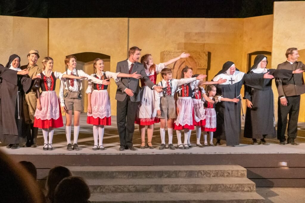 A cast of children and adults stands in a line on a stage wearing costumes for a theatrical performance.