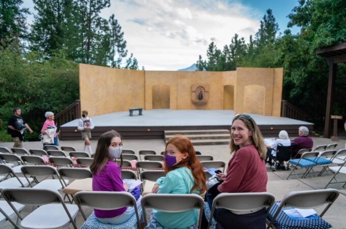 A group of people, some wearing masks, seated in a park or outdoors area with a stage in the background.