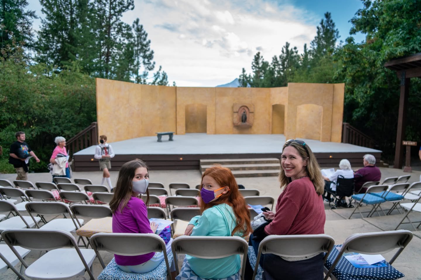 A group of people, some wearing masks, seated in a park or outdoors area with a stage in the background.