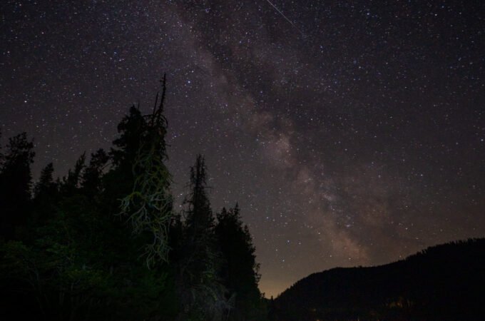 The Milky Way arches over a nighttime camping scene with people seated in a clearing.
