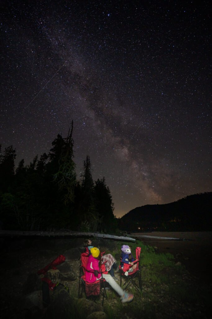 A night sky full of stars and the Milky Way above silhouetted people in camping chairs near a body of water.