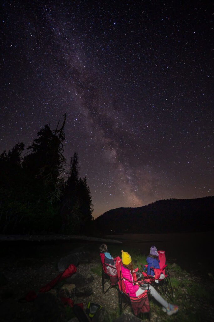 Two people stargazing under a starry night sky with the Milky Way visible. Colorful jackets and backpacks are visible in a dark outdoor setting.