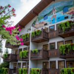 A hotel building features a vibrant beach mural and wooden balconies overflowing with greenery. Pink bougainvillea blooms in the foreground against a blue sky.
