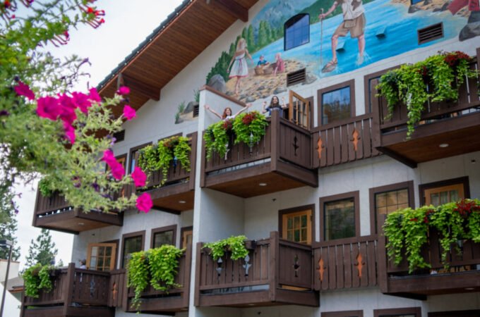 A hotel building features a vibrant beach mural and wooden balconies overflowing with greenery. Pink bougainvillea blooms in the foreground against a blue sky.