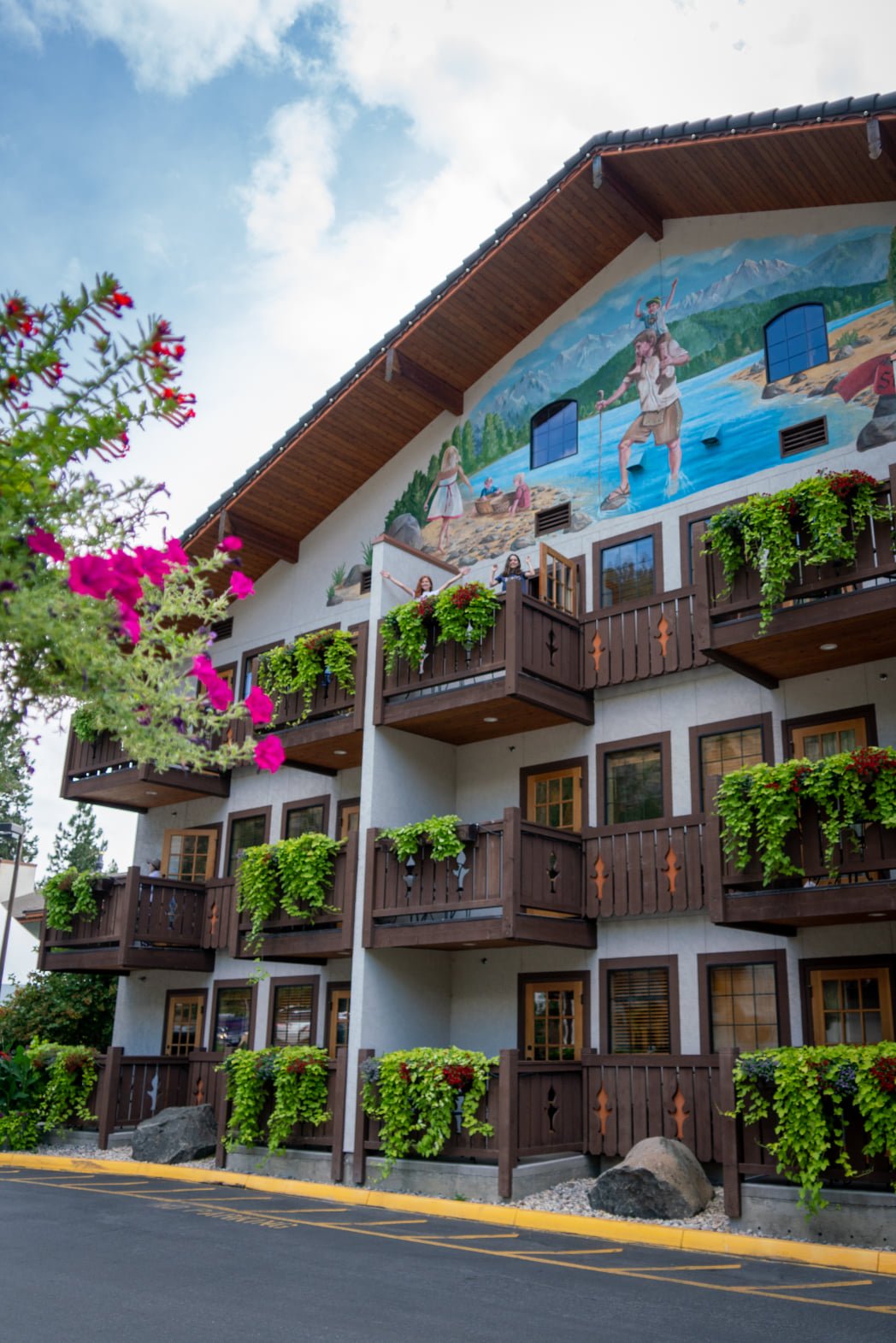 A hotel building features a vibrant beach mural and wooden balconies overflowing with greenery. Pink bougainvillea blooms in the foreground against a blue sky.