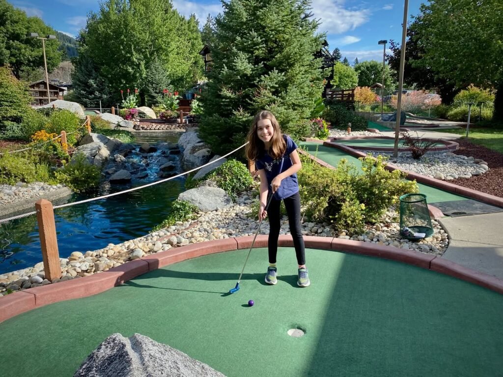 A person playing miniature golf on a scenic course surrounded by lush greenery and a stream.