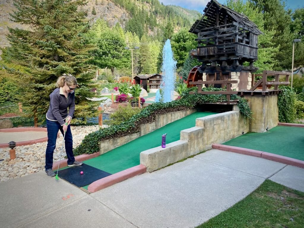 A person focuses on a put near a rustic wooden building and a fountain at a mini-golf course set against mountain scenery.