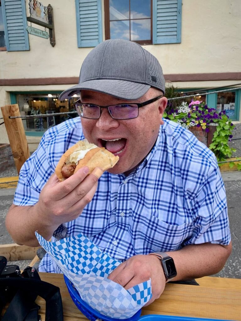 A person takes a large bite of a hot dog at an outdoor cafe.