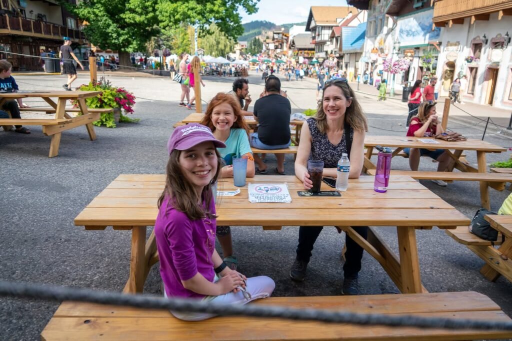 A family relaxes at a wooden picnic table in a bustling European town square. The background features traditional architecture and lively pedestrian activity.