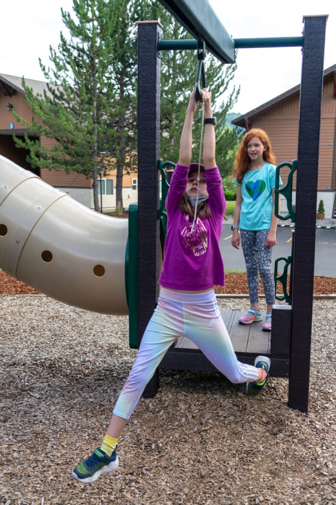 Two children playing on playground equipment with a slide in the background. One is hanging from a bar while another stands nearby.