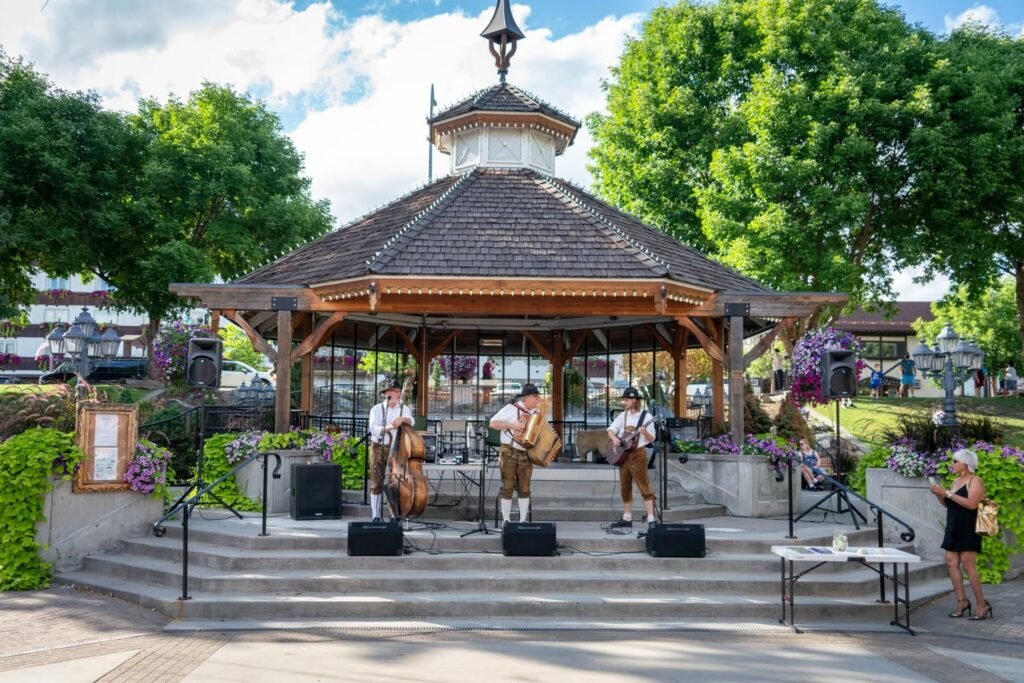Folk musicians perform on an outdoor bandstand surrounded by trees and a lush garden in the Pacific Northwest.