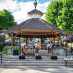 Folk musicians perform on an outdoor bandstand surrounded by trees and a lush garden in the Pacific Northwest.