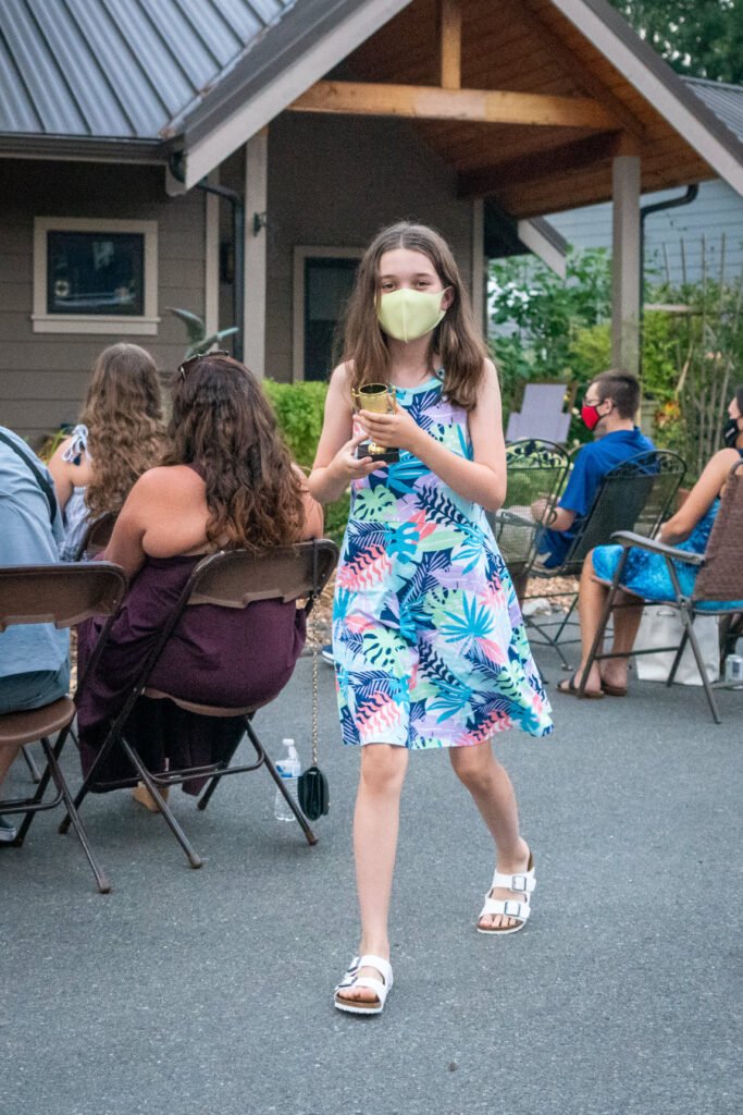 People gathered outdoors in front of a rustic cabin with a metal roof. A person in a floral dress stands near the group on the paved area.