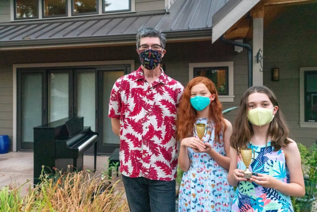 Three people wearing masks stand together outside a house, two holding trophies.