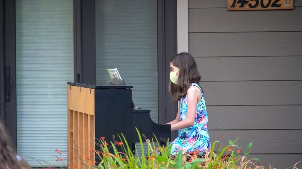 A young girl in a colorful dress plays an upright piano outside a house with a garden.