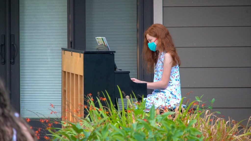 A person wearing a mask playing a piano outside a modern building with lush greenery in the foreground.