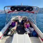 Slidewaters A group enjoying a day on a white speedboat with red accents, cruising on calm blue water with mountains in the distance.