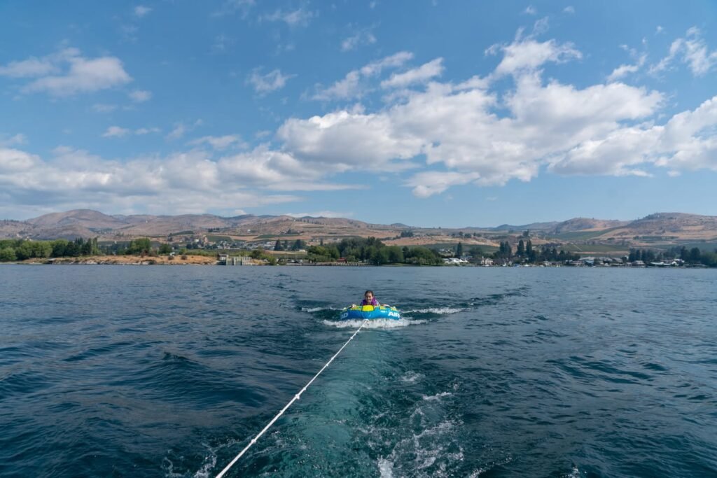 A person tubes behind a boat on a sparkling lake against a beautiful mountain backdrop under a bright blue sky. Scattered white clouds complete the scenic view from a boat.