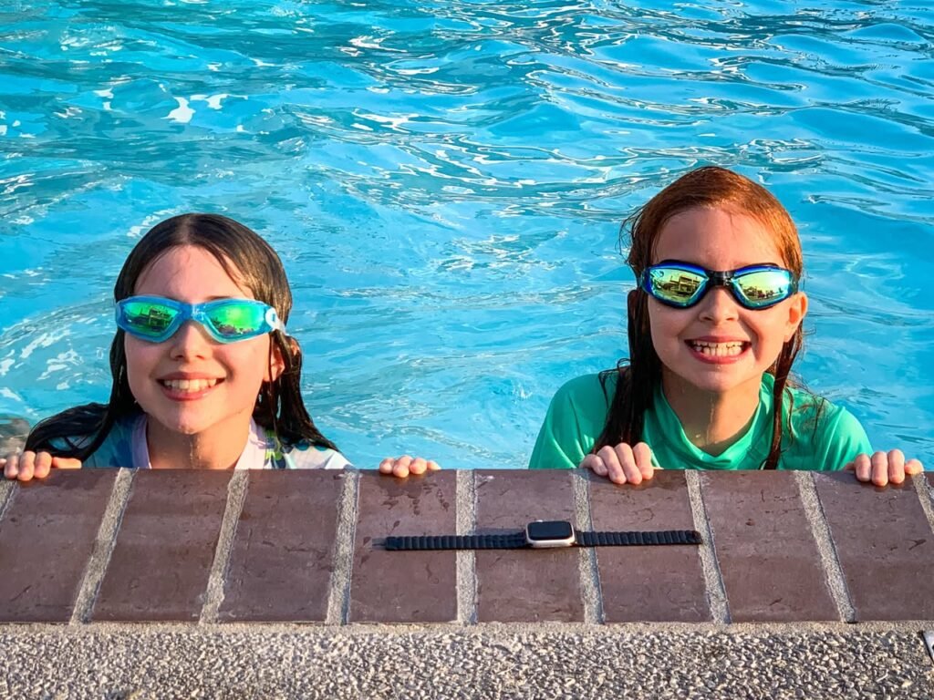 Two smiling children wearing swimming goggles peek over the edge of a bright blue pool. A black smart watch rests on the poolside brick ledge.