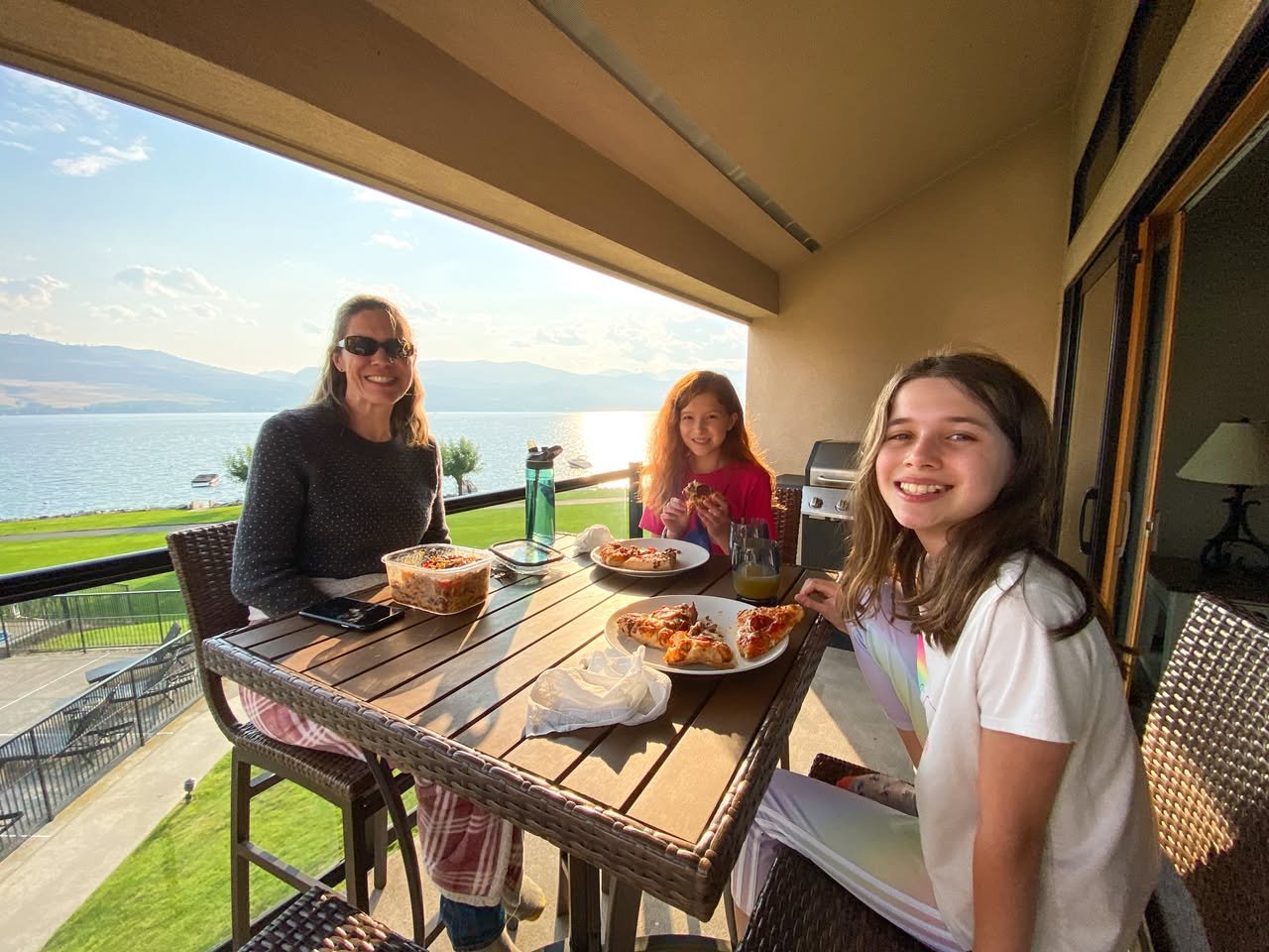 Lake Chelan Vacation Home A group enjoys a meal on a balcony overlooking a scenic lake with distant mountains. The wooden table is set with plates of food and drinks under a covered overhang.
