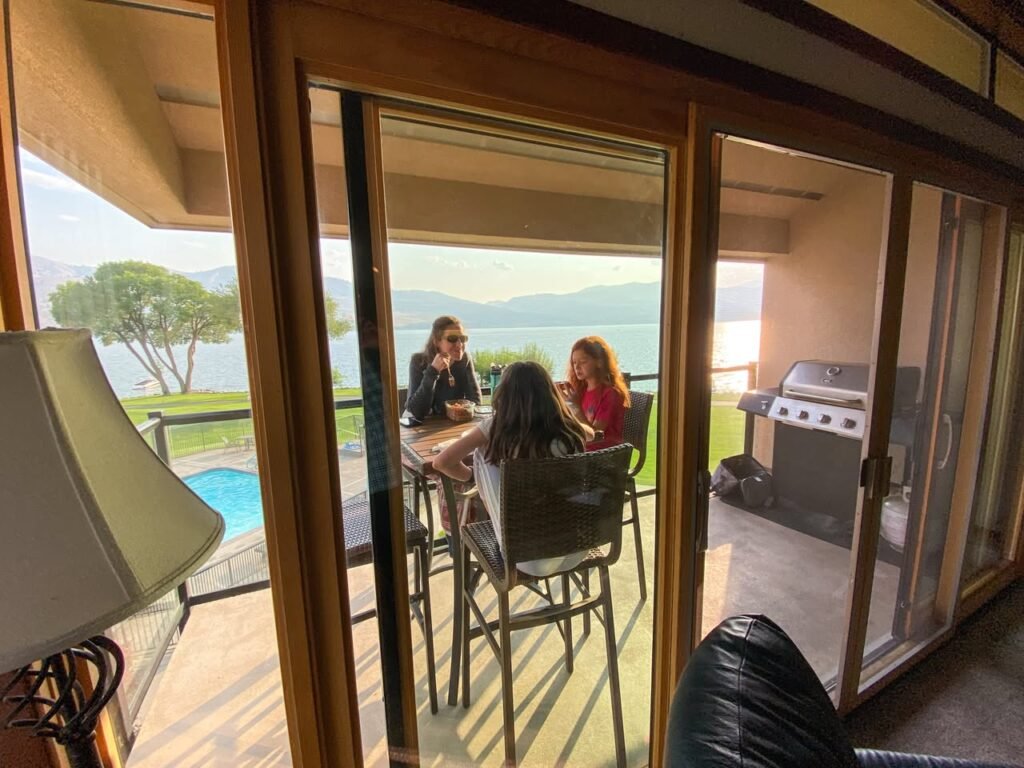 A view from inside a home looking through sliding glass doors to an outdoor deck where people are dining. A pool and distant lake are visible in the background.