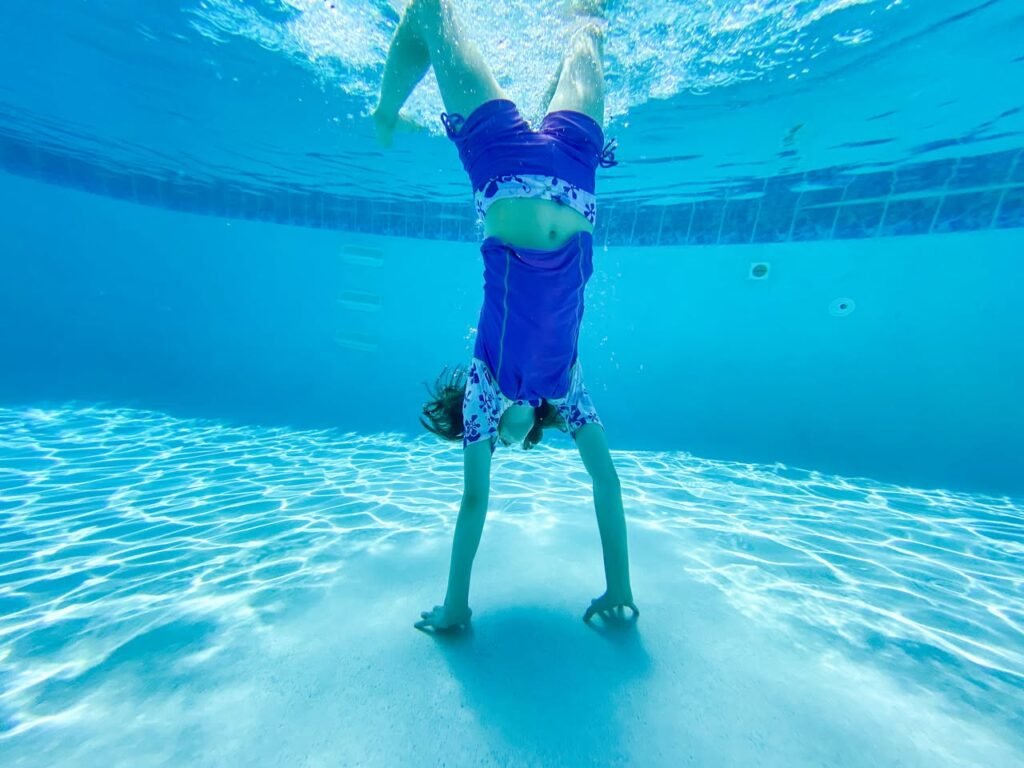 A person performing a handstand underwater in a blue pool with light patterns on the floor. The image captures the unique perspective of submerged swimming.