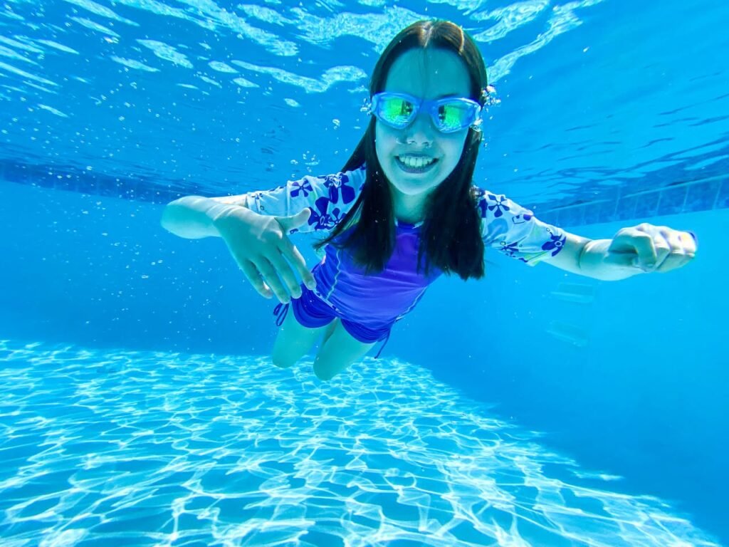 A person in a rash guard and swim goggles smiles brightly while swimming underwater in a pool. The water features sparkling caustic light patterns.