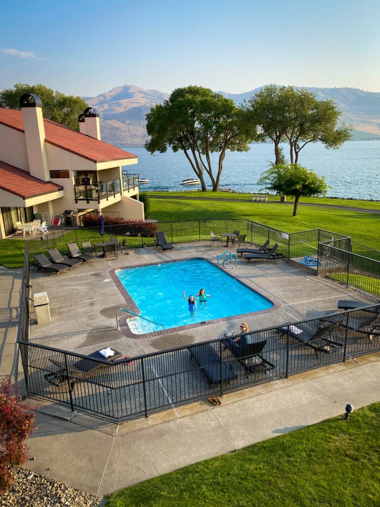A bright swimming pool with loungers overlooks a beautiful body of water and distant mountains under a clear blue sky.