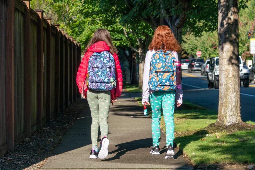 Two friends walking with colorful backpacks on a sunny sidewalk.