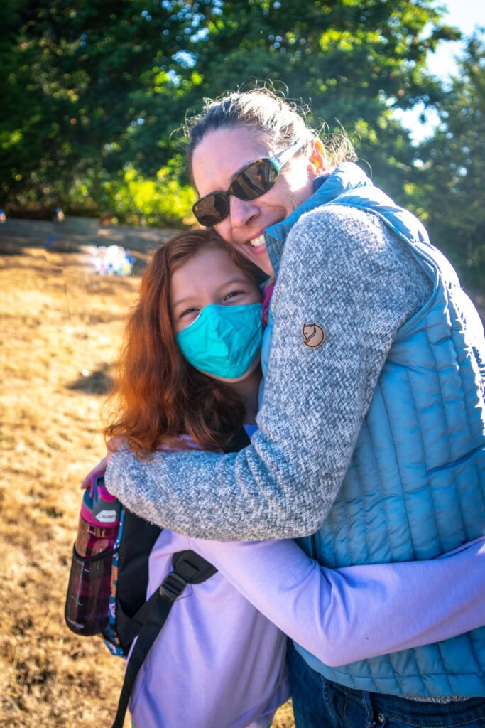 Two people hugging outdoors, both wearing face masks, with dry grass and trees in the background.