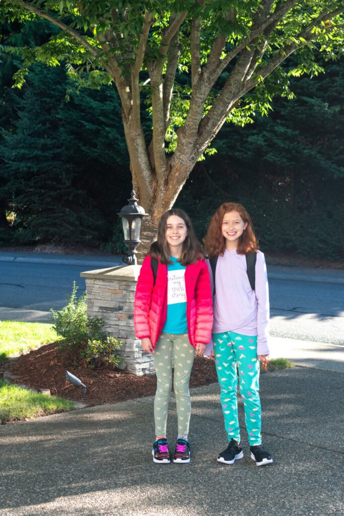 Two children pose on a quiet street in front of a large, leafy tree and a stone pillar with a lantern.