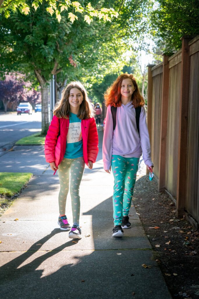 Two children walking side-by-side on a sunny sidewalk lined with lush green trees. The full photo includes close-up sections of their outfits and the leafy branches above.