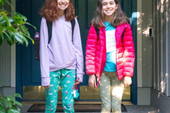 Two girls standing on the porch steps in front of a dark blue door, wearing colorful outfits and backpacks.